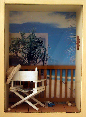 A one-twelfth scale modern miniature beach balcony scene in a box frame On balcony is a white director's chair with a woven hat on the back, a magazine on the seat, and a pair of jandals and a glass on the floor. The balcony overlooks the sea and some cabbage trees.
