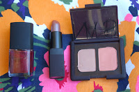 picture of Nars nail polish in a shimmery burgundy red color, lipstick in a pink nude, and eyeshadow in bohemian gold on a blue, yellow, pink and orange flowered background
